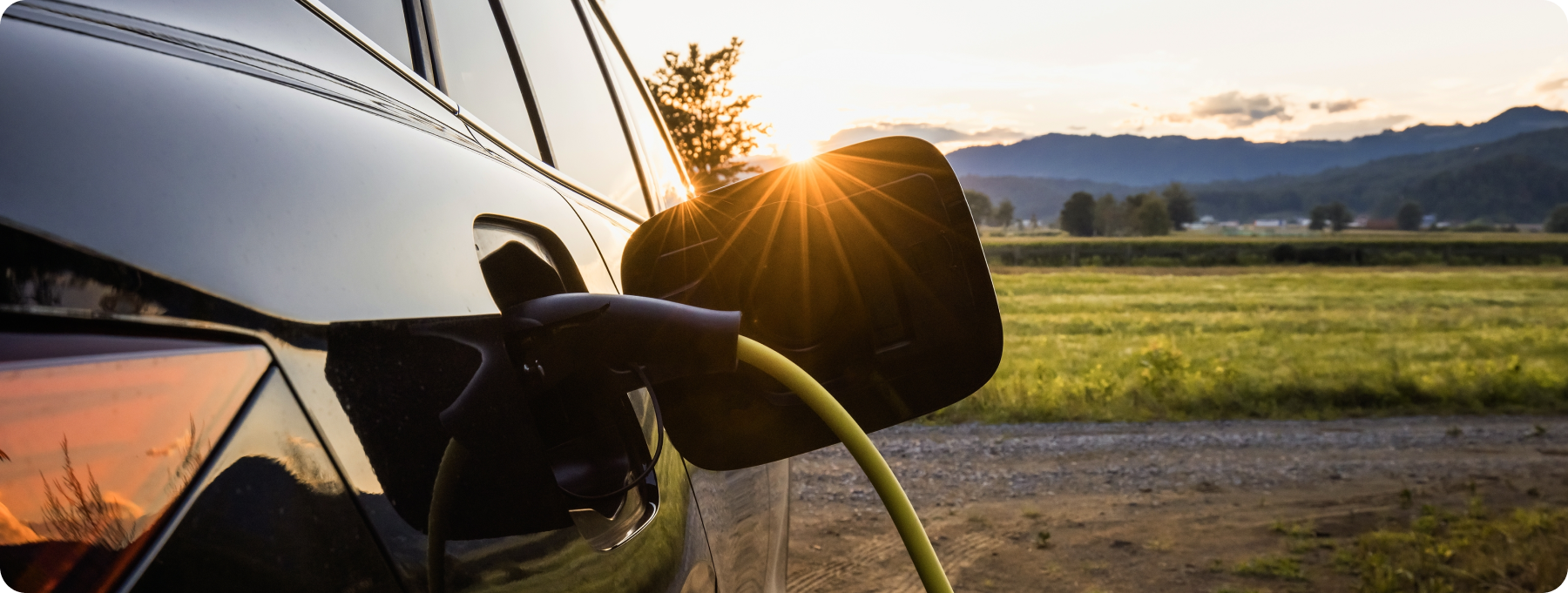 La photographie montre une voiture électrique branchée à un câble de recharge, avec le soleil couchant qui brille au-dessus du retroviseur, en arrière plan. L'image suggère d'utiliser l'énergie solaire pour déployer la mobilité électrique.